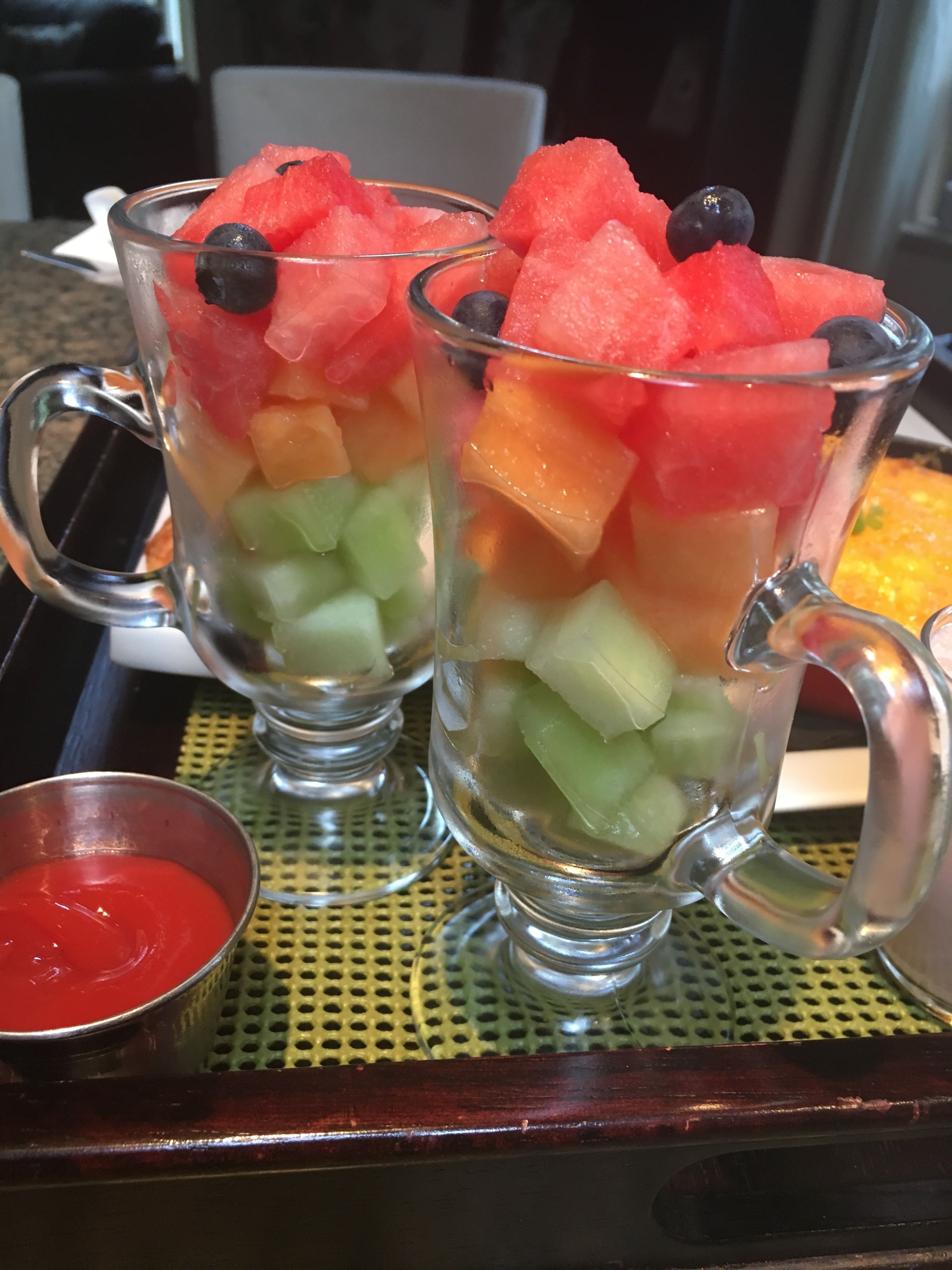 Two clear glasses filled with layered fruit and a small metal cup of red sauce on a tray.