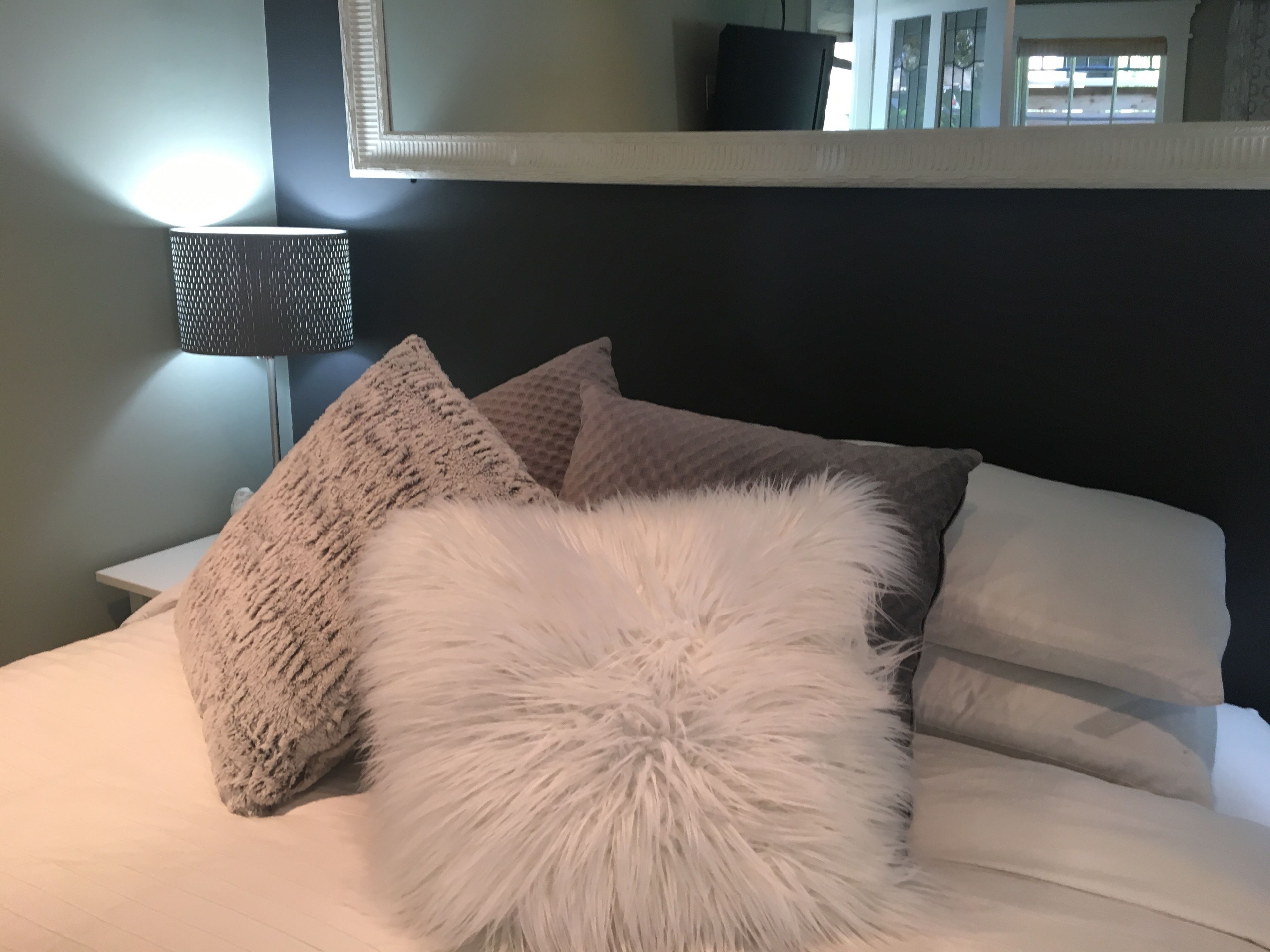 A bed with a white fluffy pillow, two textured pillows, and a lamp on a bedside table.