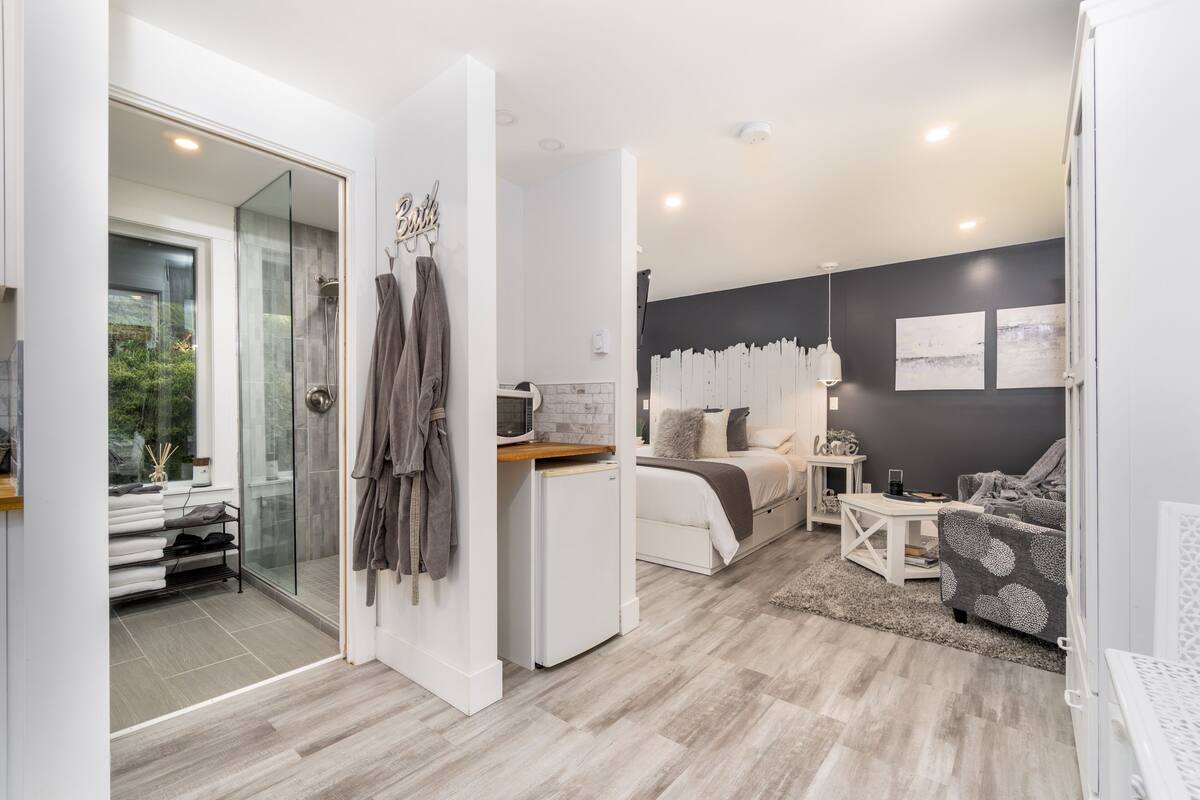 Une chambre moderne avec une douche en verre, un lit doté d’une tête de lit blanche, un petit bureau blanc et un fauteuil à motifs.
