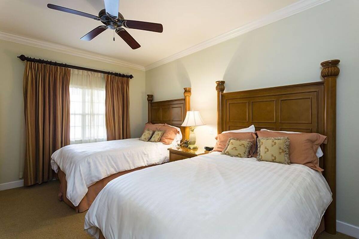Habitación de hotel con dos camas, ventilador de techo, mesita de noche con lámpara y cortinas.