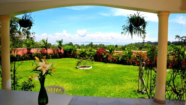 A covered patio with a view of a well-maintained lawn and garden. - Victoria Lake View Guest House & Safaris (Entebbe)