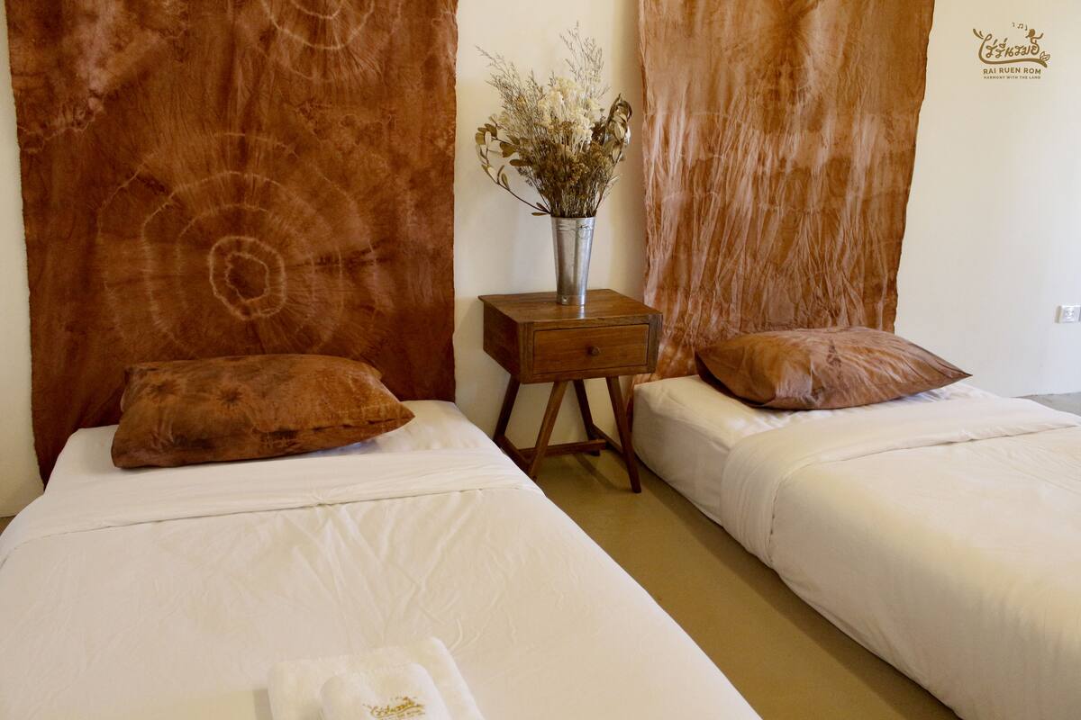 A hotel room with two beds, a wooden nightstand with a vase of flowers, and brown curtains.