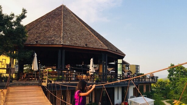 A wooden bridge leading to a building with a thatched roof, outdoor seating, and a view of the surrounding greenery. - Rai Ruen Rom Organic Farm (Thoeng)