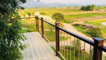 Um deck de madeira com guarda-corpo de metal, com vista para uma paisagem rural com campos e uma cordilheira ao longe.
