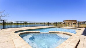 A hot tub with a curved design, surrounded by a black metal fence, overlooking a body of water and a building in the background.