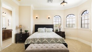 A bedroom with a large bed, two bedside tables, a nightstand, a window with blinds, and a door leading to a bathroom.