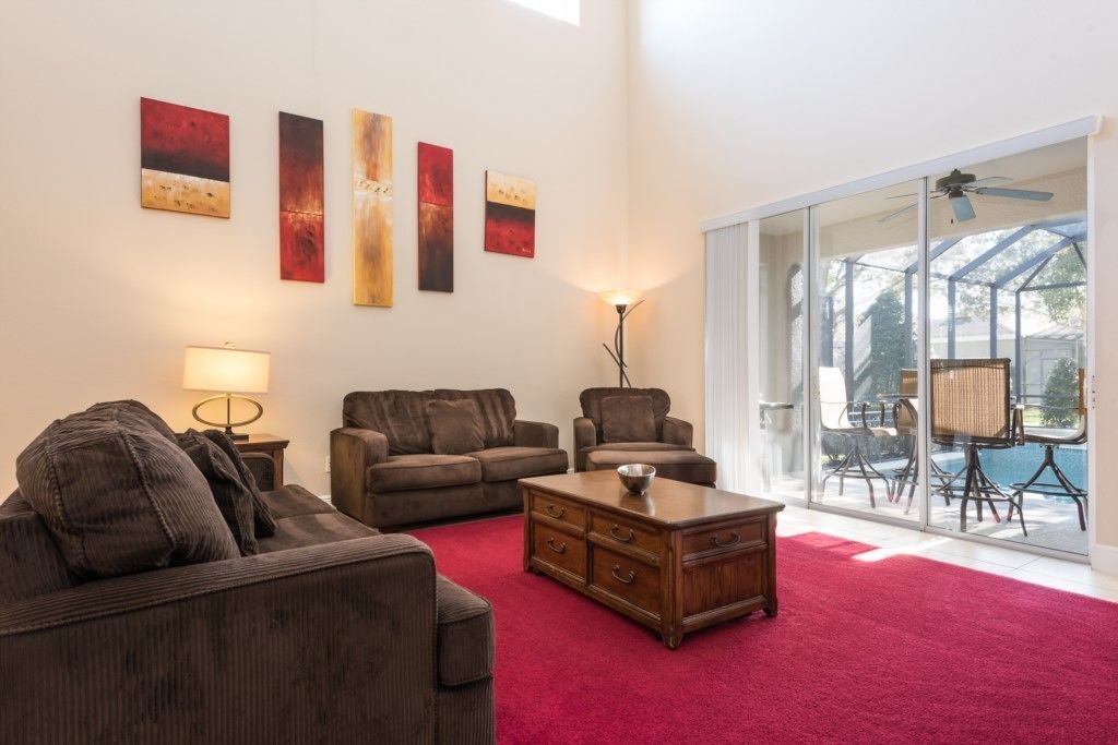 Een woonkamer met een rood tapijt, twee banken, een salontafel en een lamp. Aan de muur hangen abstracte schilderijen en er is een glazen deur die leidt naar een buitenruimte met een zwembad.