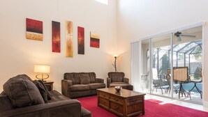 Ett vardagsrum med en röd matta, två soffor, ett soffbord och en lampa. På väggen hänger abstrakta målningar, och en glasdörr leder ut till ett utomhusområde med en pool.
