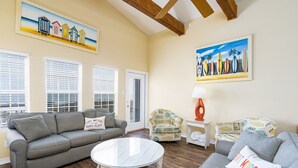 A living room with a grey sofa, a round glass coffee table, and a patterned armchair. There are two windows with blinds, a painting of beach huts, and a painting of surfboards.