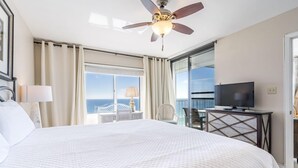 A hotel room with a large bed, a ceiling fan, a TV, a balcony with a view of the ocean, and a dining area with a table and chairs.