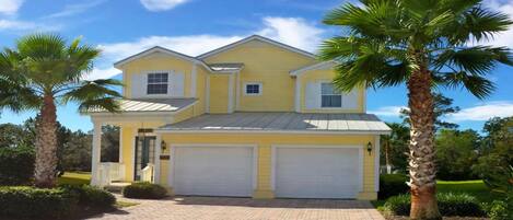 A two-story yellow house with a garage, surrounded by palm trees and a well-maintained lawn.