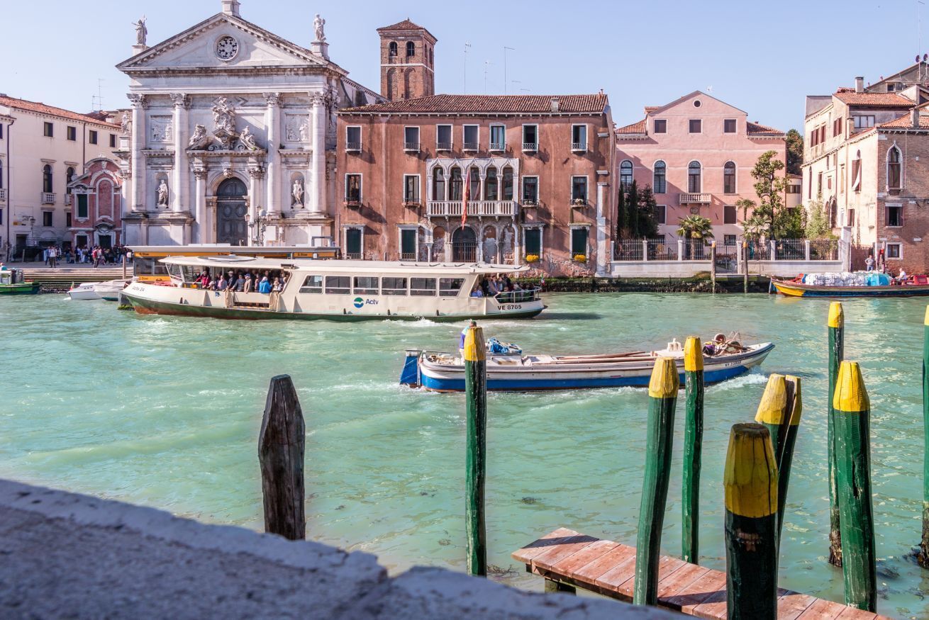 Um canal com barcos, um edifício histórico com uma torre de relógio e uma fileira de edifícios venezianos tradicionais.