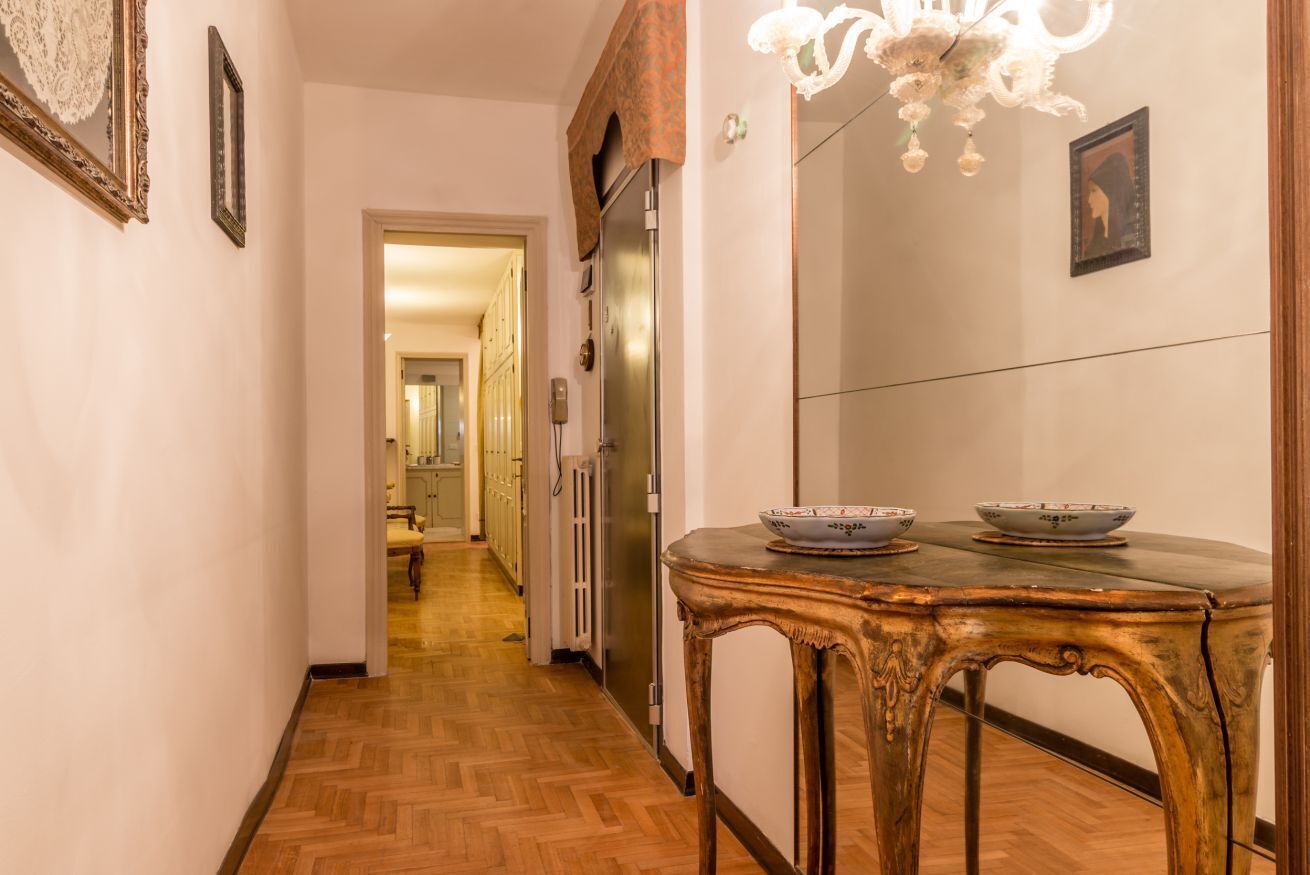 A hallway with a wooden floor, a vintage table with two bowls, a chandelier, and framed pictures on the walls.