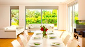 A modern dining area with a white table set for six, a white sofa, and large windows offering a view of greenery.