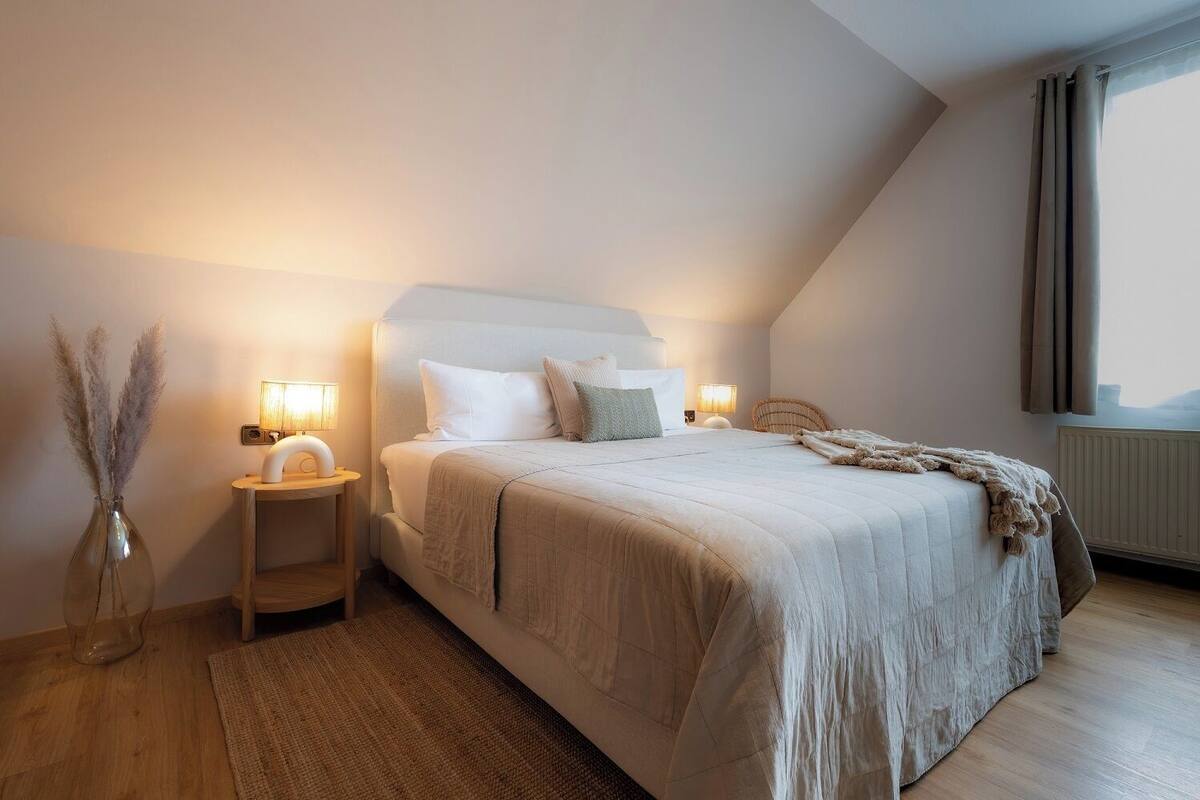 A bedroom with a bed, bedside tables, a lamp, a vase with pampas grass, and a window with curtains.