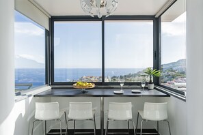 A modern kitchen with a large window, a dining table with four white chairs, and a bowl of fruit. - Caniço VI, spacious apartment with pool. (Caniço)