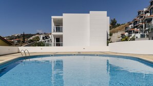 Une piscine à l’eau d’un bleu limpide, entourée d’un bâtiment blanc doté de balcons, et offrant une vue sur une colline avec des bâtiments en arrière-plan.