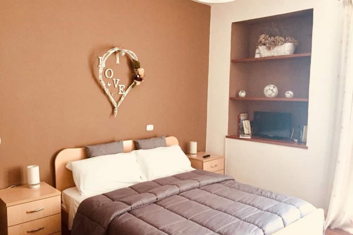 Un dormitorio con cama, mesitas de noche, una estantería con adornos y una decoración con forma de corazón en la pared.