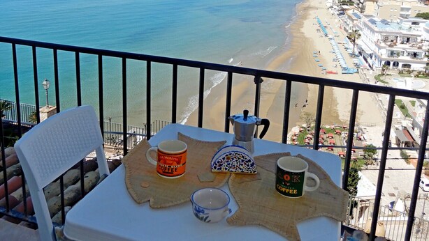 Ein Balkon mit Tisch für zwei Personen, Blick auf einen Strand mit klarem, blauem Wasser und eine Stadtlandschaft in der Ferne.
