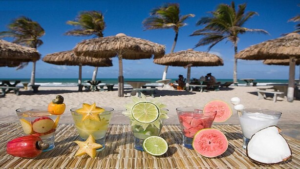 A beach setting with thatched umbrellas, a table with various fruit and drink garnishes, and a coconut on the table. - Apartamento Luxo vista mar, muito aconchegante VP803 (Fortaleza)