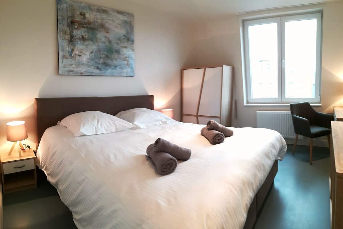 A neatly made bed with pillows and towels, a bedside lamp, a large window, and a painting on the wall.