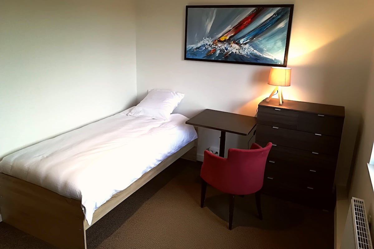 A single bed with white bedding, a small desk with a lamp, a red chair, and a dresser.