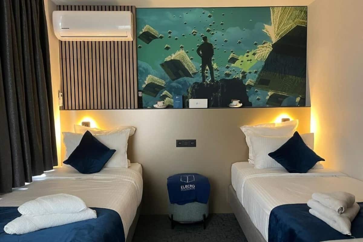 A hotel room with two beds, a large artwork above the beds, an air conditioning unit, and a small ottoman with a towel.