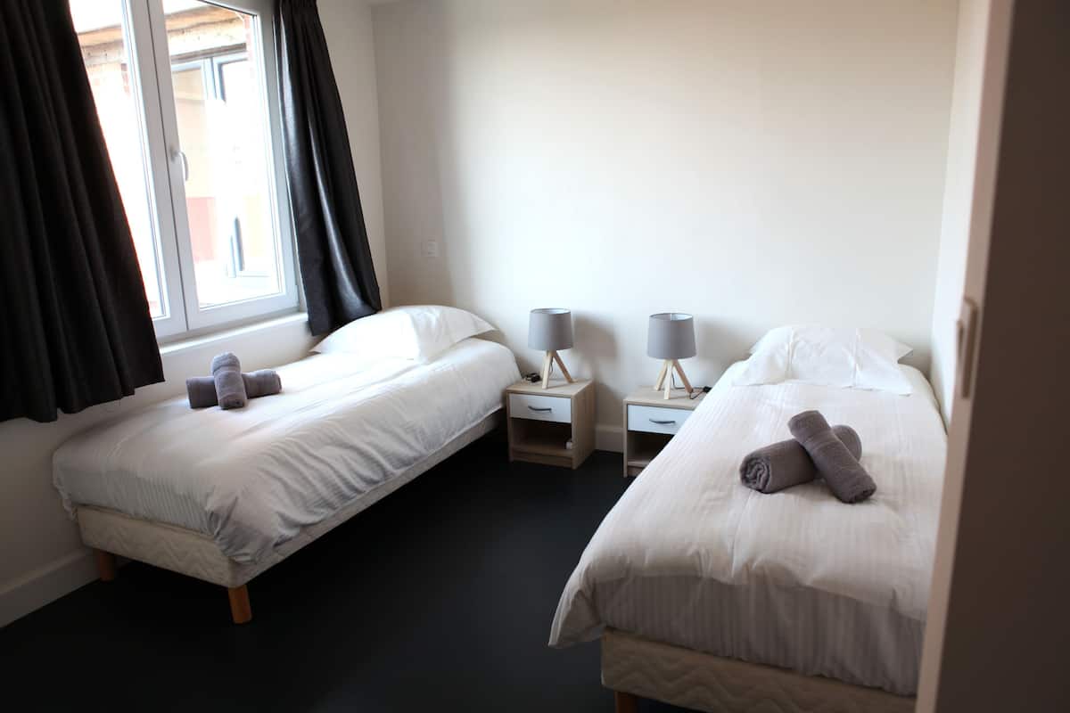 A hotel room with two single beds, bedside tables with lamps, and a window with curtains.