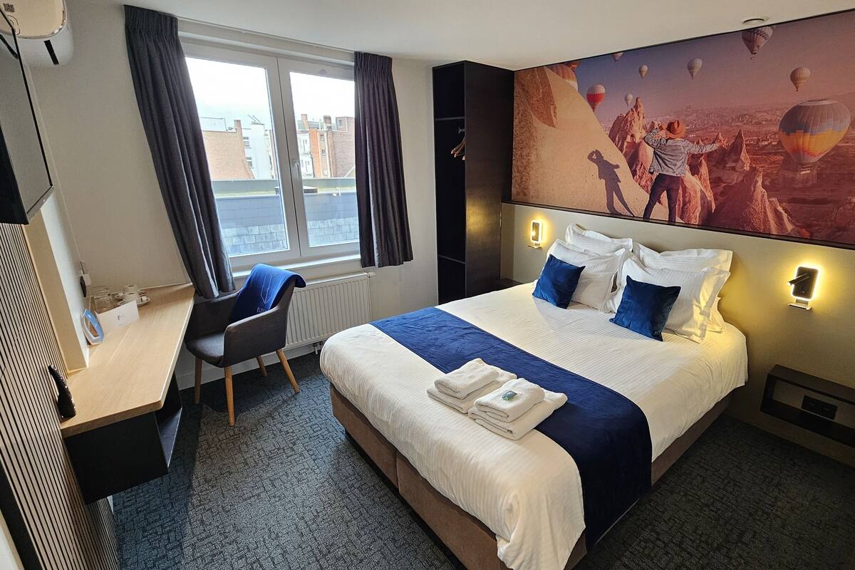 A hotel room with a large bed, a desk, a chair, and a view of the city through the window.