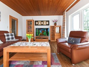 Ein Wohnzimmer mit Holzbalkendecke, einem Fernseher, einer Anrichte und zwei Ledersofas.