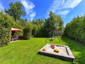 Ein Garten mit Sandkasten, Schaukel und einer Terrasse mit einem roten Sonnenschirm.