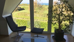 Habitación moderna con un ventanal grande que ofrece vistas a un viñedo y al mar. Se observa una silla negra y una planta en maceta.