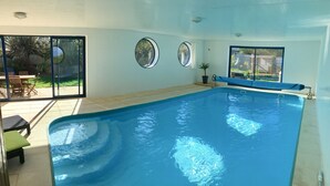Una piscina con plataforma de baldosas, una tumbona y un ventanal de vidrio que da a un espacio exterior con patio y sillas.