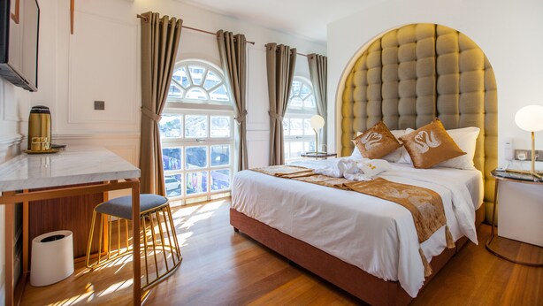 Ein Hotelzimmer mit einem großen Bett, einem Schreibtisch und einem Stuhl. Vor den Fenstern hängen Vorhänge und auf dem Nachttisch steht eine Lampe.