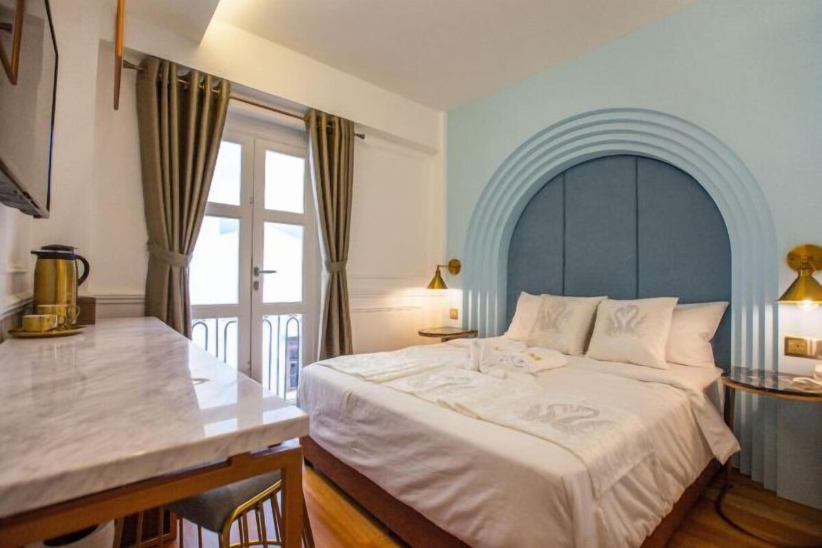 A hotel room with a bed, a table, and a chair. There is a window with curtains, a lamp, and a wall with a circular design.