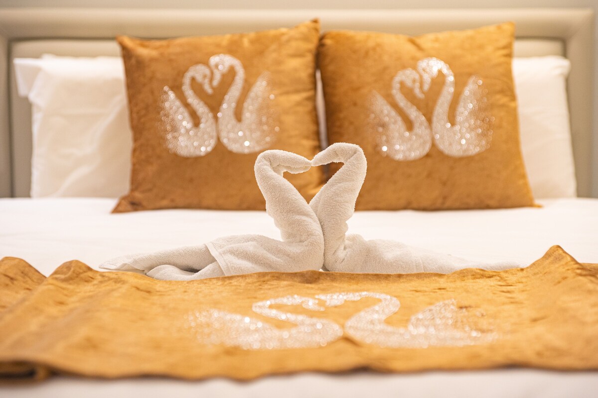 A bed with two pillows featuring swan designs, a white blanket, and a towel arranged in a heart shape.