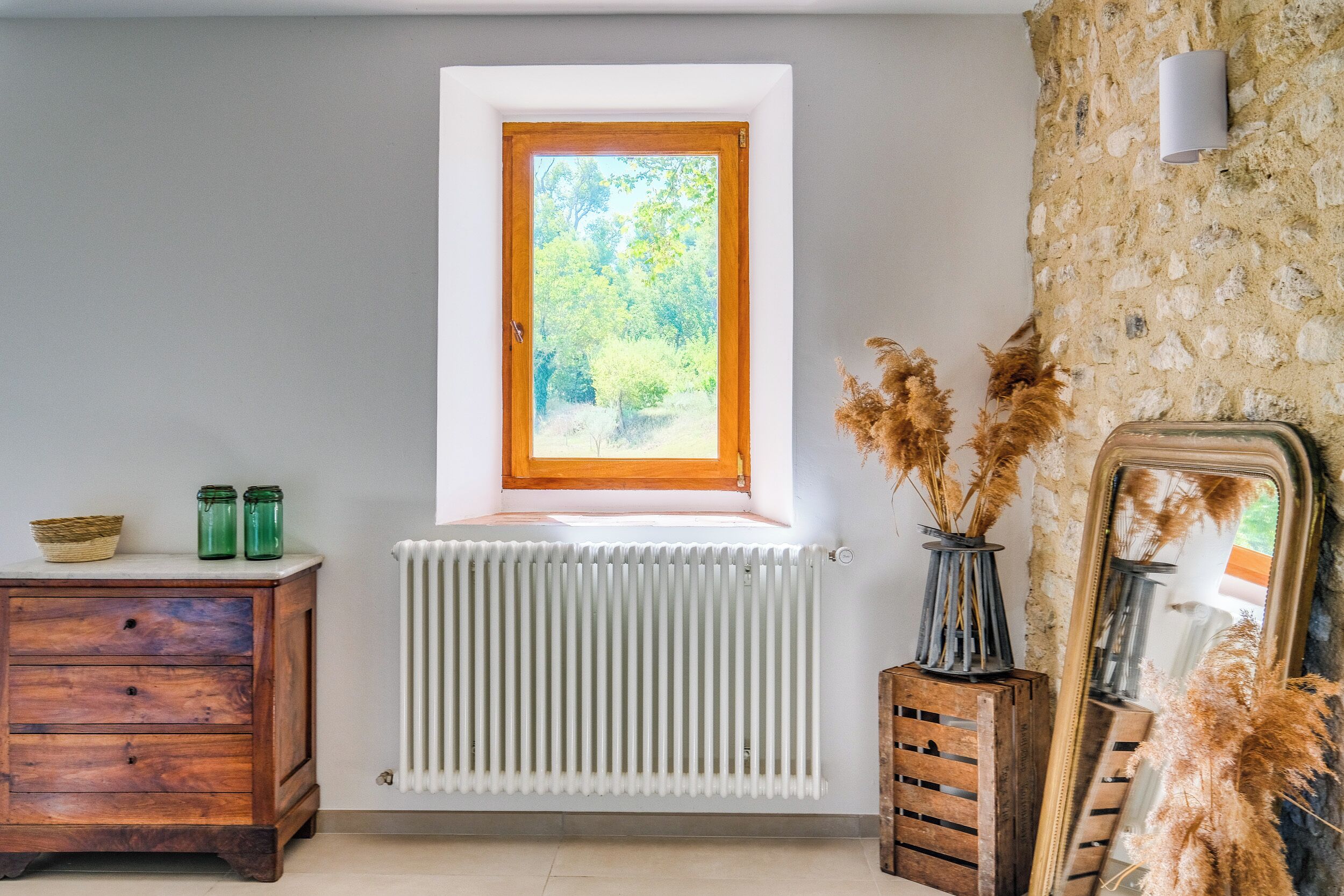 A room with a wooden dresser, a white radiator, a window with a view of greenery, and a mirror with pampas grass.
