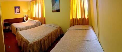 A hotel room with two single beds, a desk, a chair, and a wall-mounted lamp.