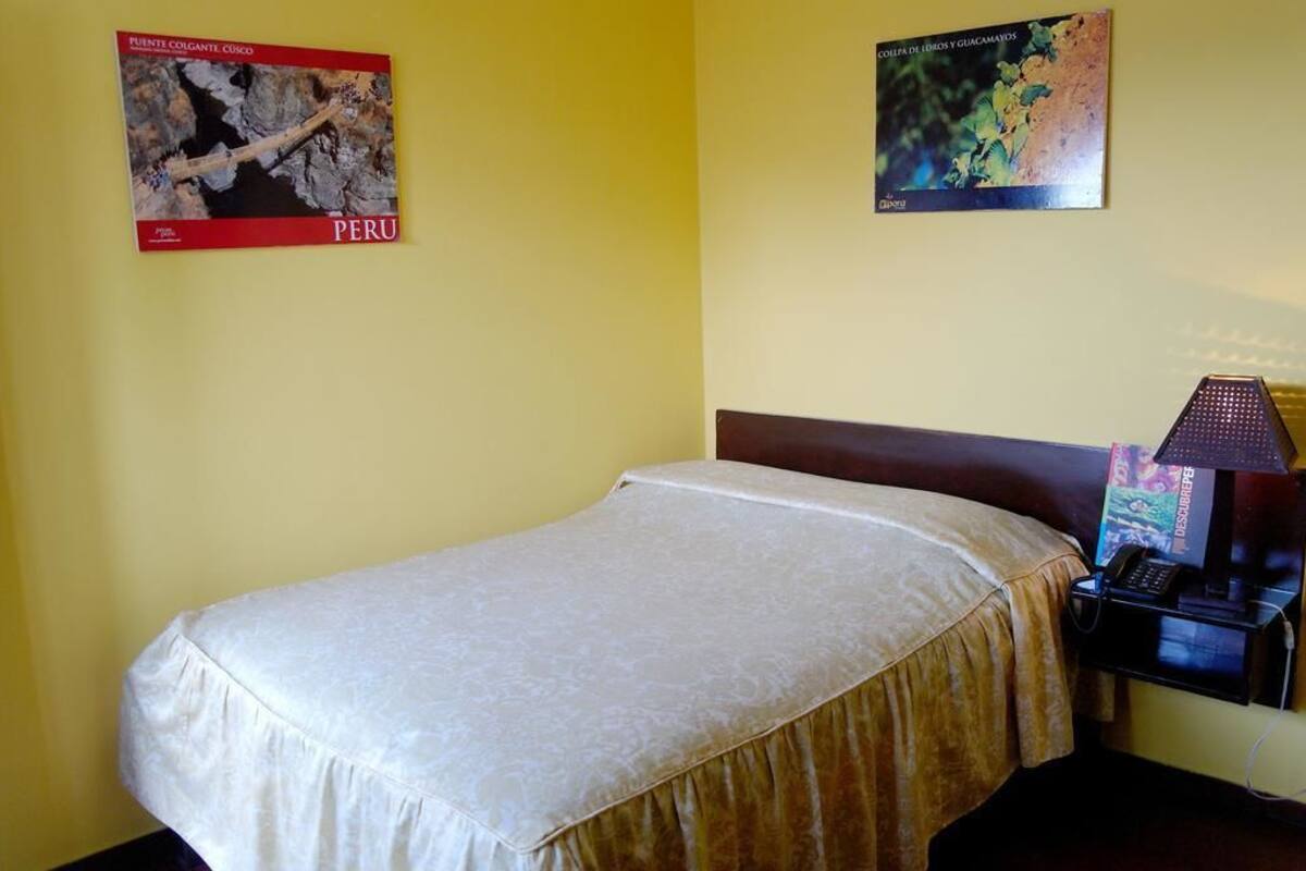 A hotel room with a single bed, a nightstand with a lamp, and two wall-mounted posters of Peru.