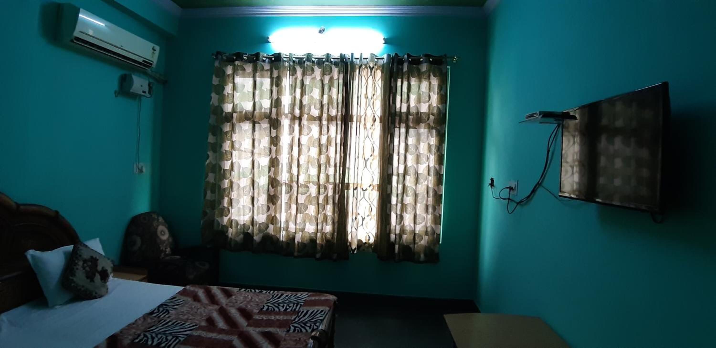 Una habitación con paredes verde azulado, una cama con ropa de cama estampada, un ventanal con cortinas estampadas y un televisor montado en la pared.
