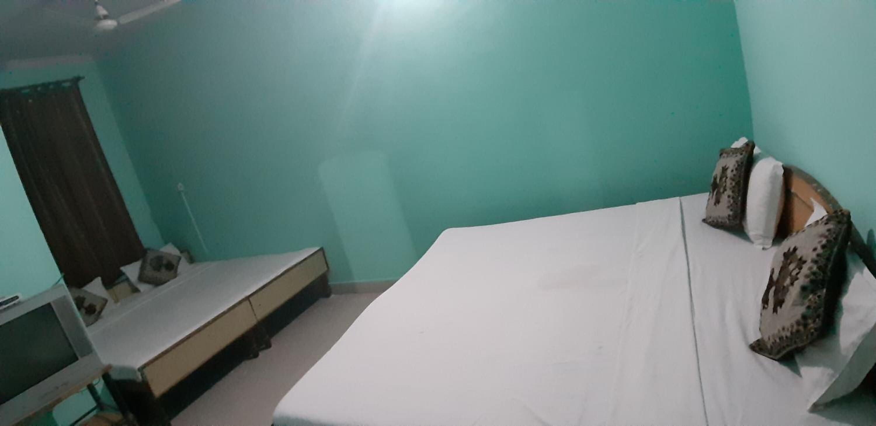 Quarto com paredes verde-água, uma cama com almofadas estampadas, uma pequena escrivaninha e uma janela com cortinas.