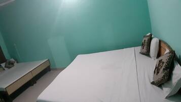 Quarto com paredes verde-água, uma cama com almofadas estampadas, uma pequena escrivaninha e uma janela com cortinas.