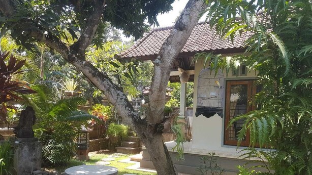 A garden with a stone table and chairs, a small statue, and a house with a tiled roof. - A Newly 1-Bedroom House with Open Kitchen Overlooking to Garden (Amed)
