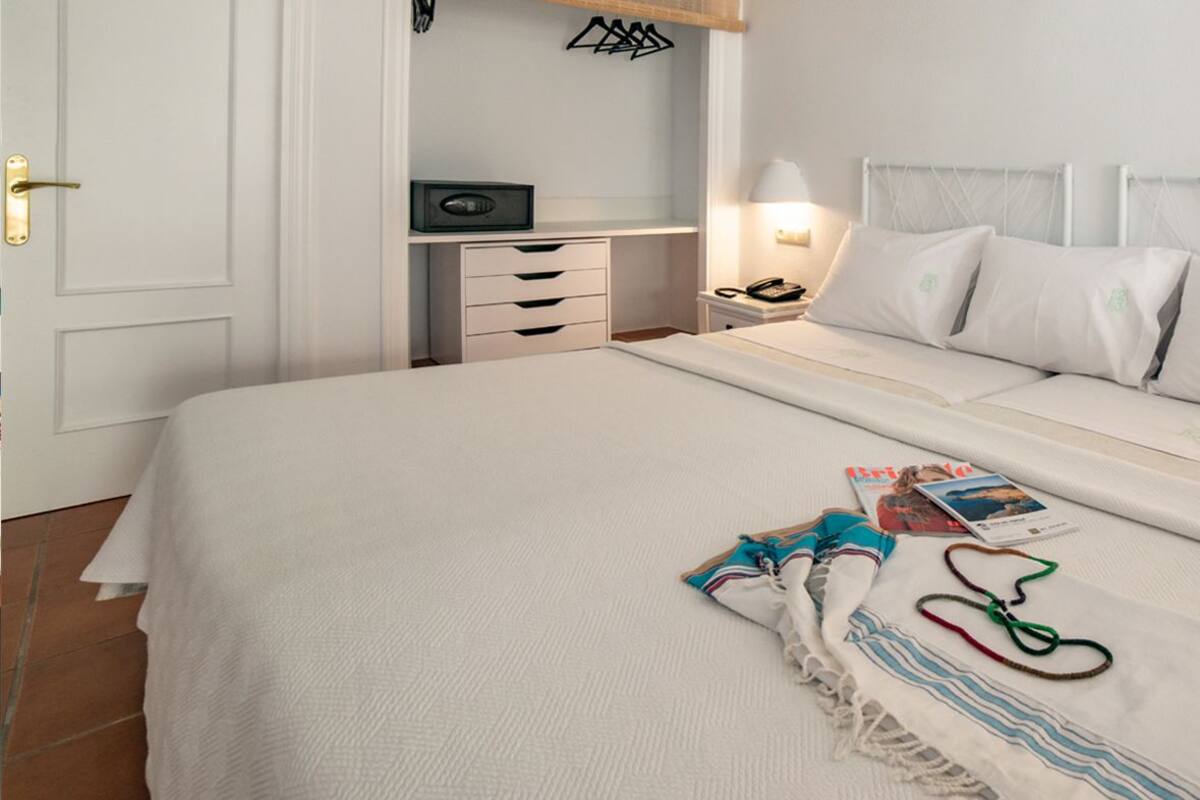 A neatly made bed with a white comforter and pillows, a bedside table with a lamp, a radio, and a magazine.
