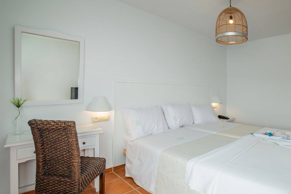 A hotel room with a bed, a desk with a chair, a bedside table with a lamp, and a hanging light fixture.