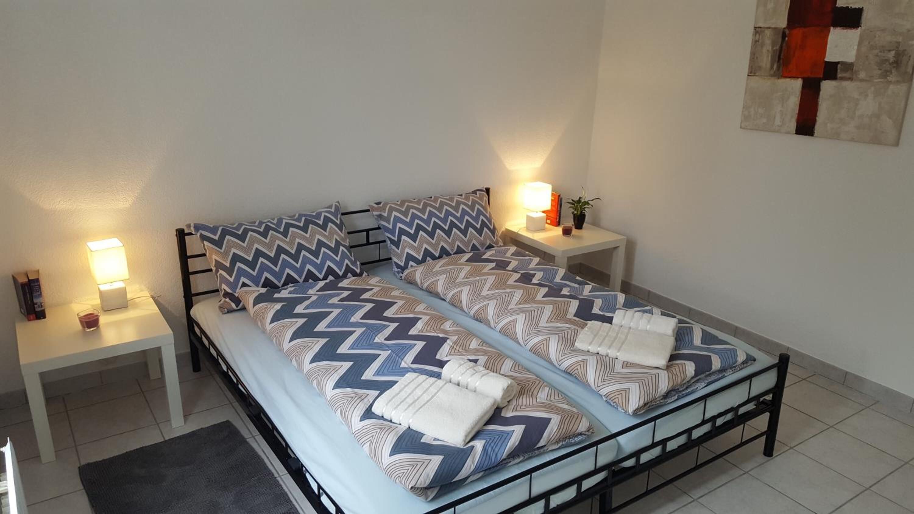 A double bed with a geometric patterned bedspread, two nightstands with lamps, a small table with a plant, and a wall art piece.