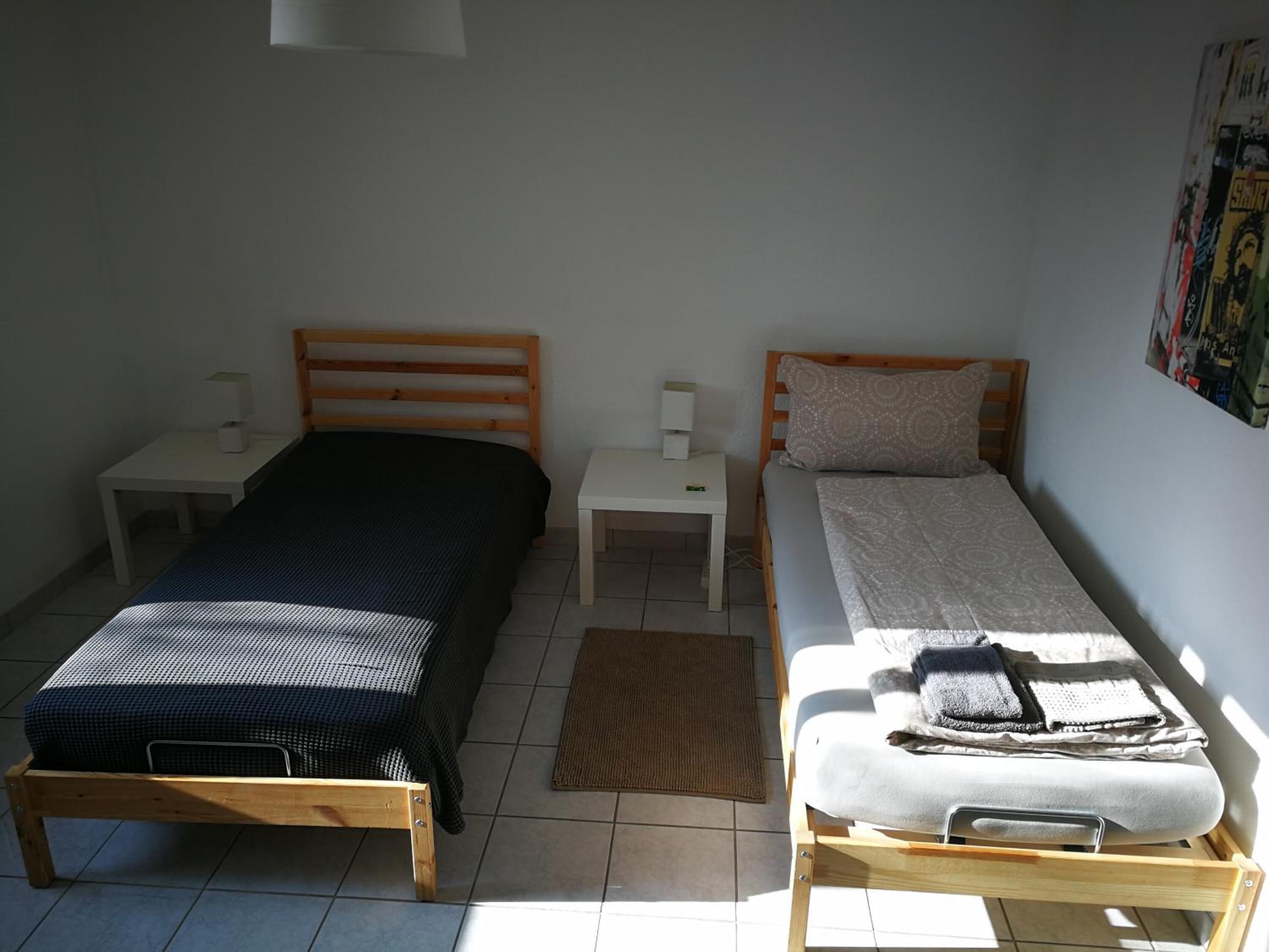 Une chambre avec deux lits simples, chacun doté d’un cadre en bois et d’une tête de lit. On y trouve des tables de chevet avec des lampes, un tapis sur le sol et un mur orné d’une affiche.