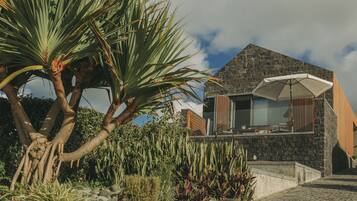 A modern house with a stone facade, a balcony with a white umbrella, and lush tropical plants.