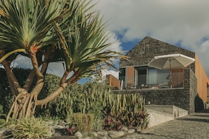 Uma casa moderna com fachada de pedra, uma varanda com guarda-sol branco e plantas tropicais exuberantes.