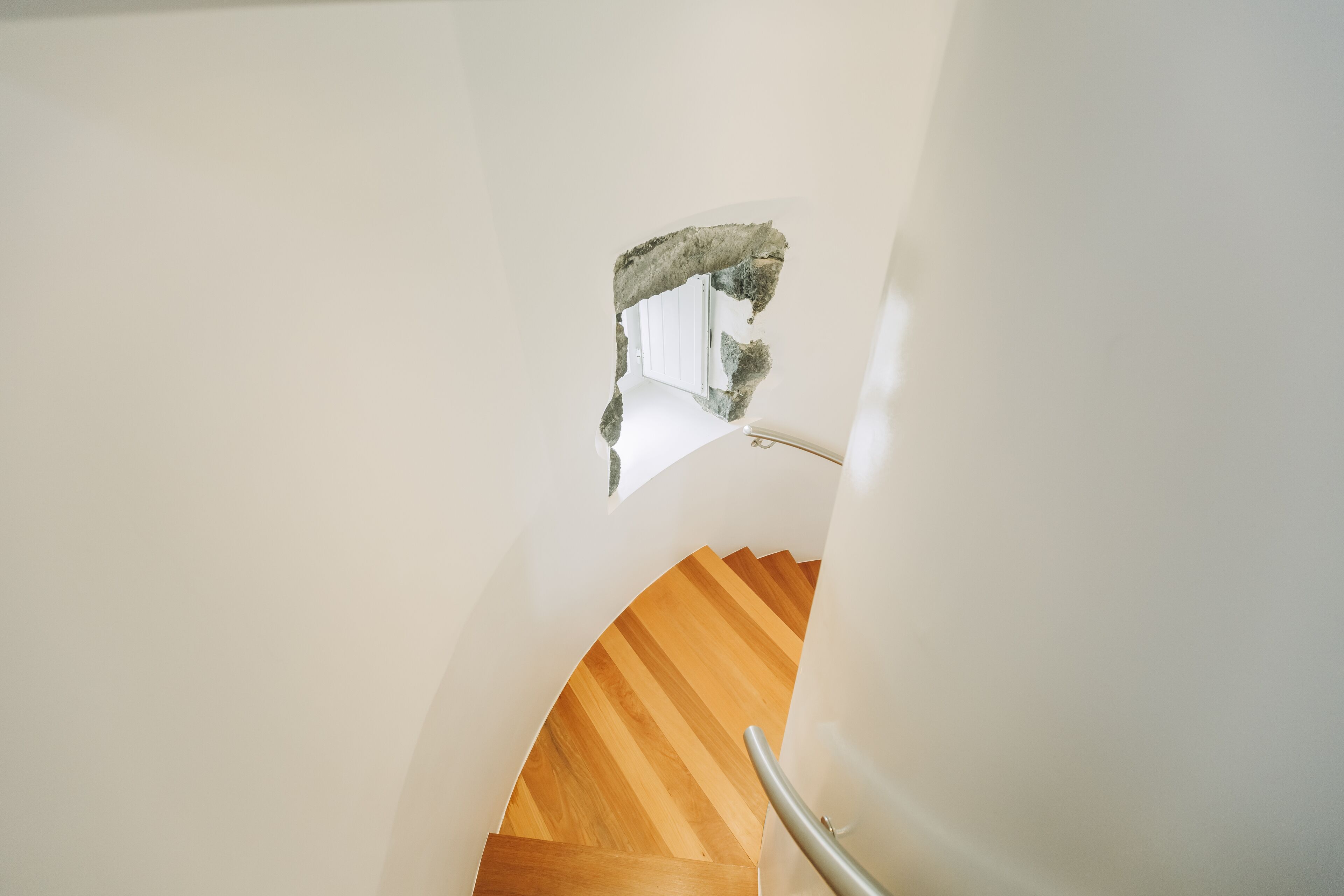 Una escalera de caracol con escalones de madera y barandilla metálica, que conduce a una habitación con paredes blancas y un ventanal pequeño.